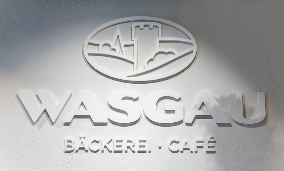 Weißes Wandschild mit dem WASGAU-Logo - einem Turm, Hügeln und Gebäuden - über dem fetten Text "WASGAU". Darunter steht in kleineren Buchstaben "BÄCKEREI - CAFÉ". Das gut beleuchtete WASGAU-Schild wirft subtile Schatten und wirkt einladend.