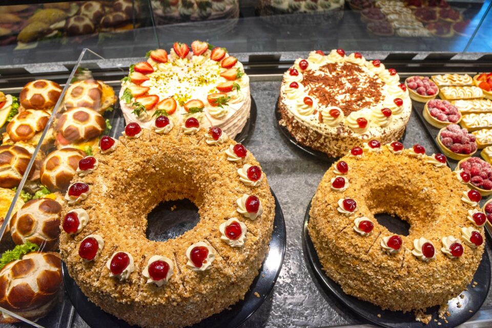 Eine Auslage der WASGAU Bäckerei mit verschiedenen Kuchen, darunter zwei ringförmige Kuchen mit Kirschen und Nüssen, ein runder Kuchen mit Erdbeeren und anderes Gebäck und Mini-Torten.