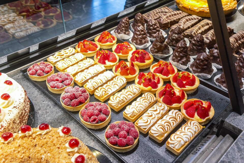 Eine Vitrine der WASGAU Bäckerei, gefüllt mit verschiedenen Backwaren, darunter Obstkuchen mit Himbeeren und Erdbeeren, Zitronen-Baiser-Torte, mit Schokolade überzogene Leckereien und ein Käsekuchen mit Kirschen.