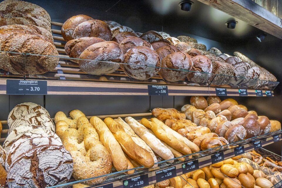 In den Regalen einer WASGAU Bäckerei gibt es eine Vielzahl von frischem Brot, darunter runde, dunkle Brote im obersten Regal und hellere Baguettes und Brötchen darunter, jeweils mit sichtbaren Preisschildern.