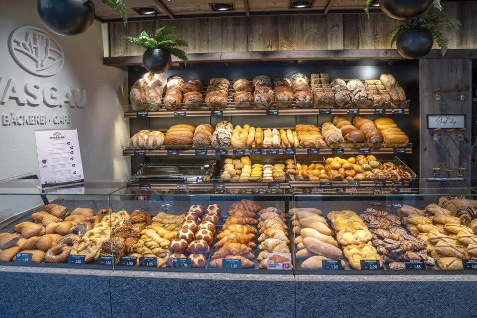 Die Auslage einer WASGAU Bäckerei mit einer Vielzahl von Brotsorten und Gebäck, ordentlich in Regalen und hinter einer Glastheke angeordnet. Schilder und Preise sind sichtbar, Brote, Brötchen und süße Backwaren gibt es in Hülle und Fülle.