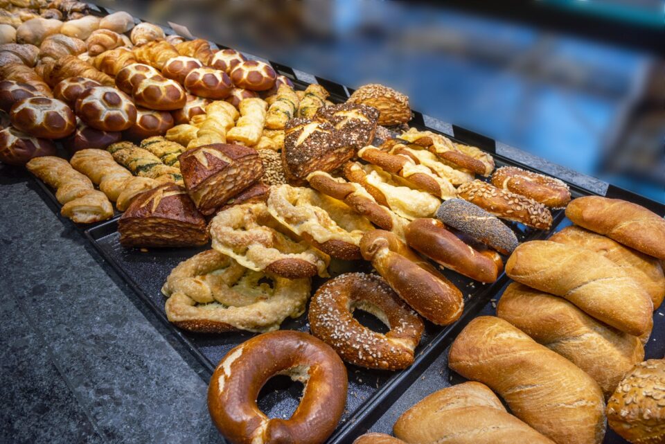 Die Auslage der WASGAU Bäckerei ist gefüllt mit verschiedenen Arten von frischem Brot und Gebäck, darunter geflochtene Brote, Brezeln, Körnerbrötchen und Baguettes, die alle fein säuberlich auf dunklen Tabletts angeordnet sind.