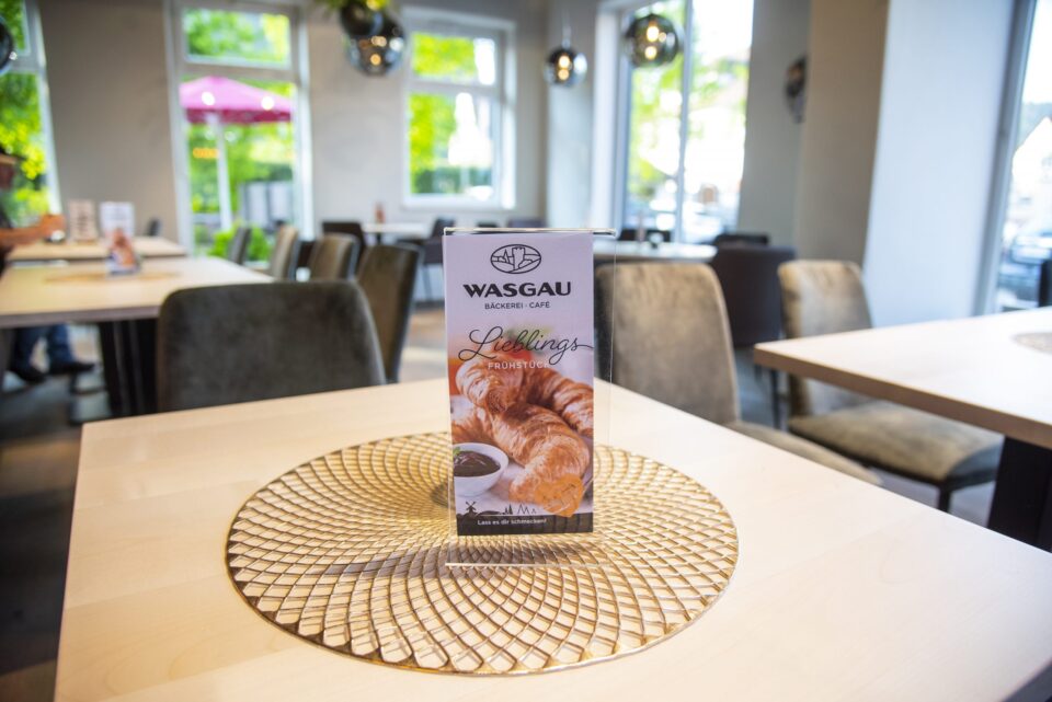 Ein Tischaufsteller in einem WASGAU Café zeigt eine Speisekarte mit Croissants und einer Tasse Kaffee. Der helle Holztisch mit einem dekorativen goldenen Tischset steht in einem hellen Innenraum mit großen Fenstern.