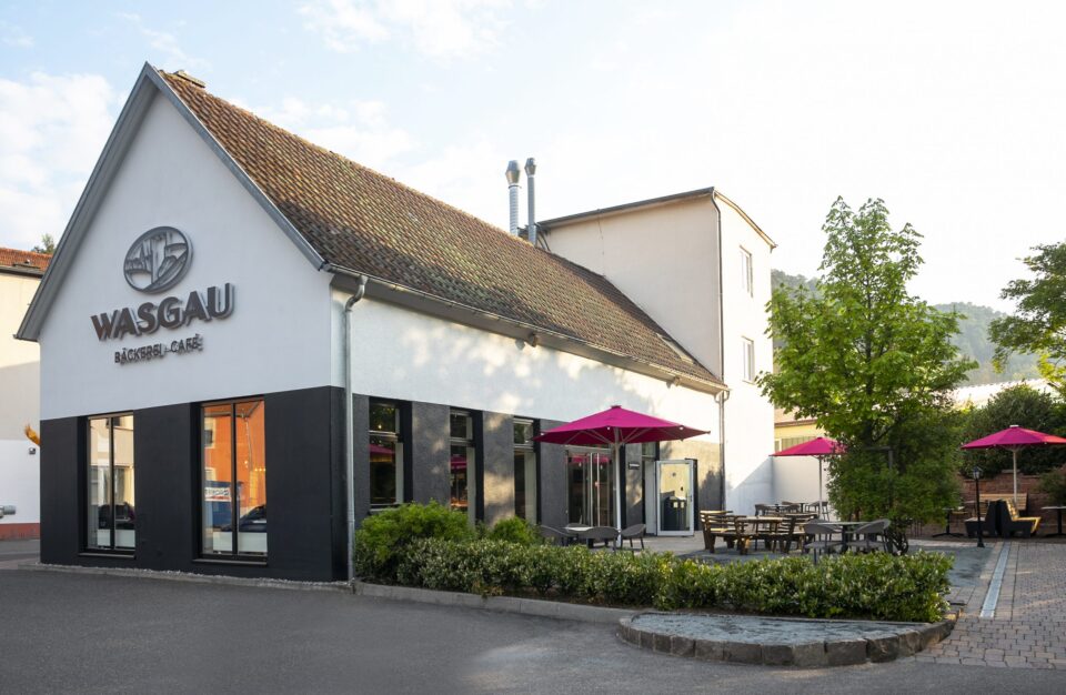 Ein modernes Gebäude mit einem WASGAU Bäcker-Café-Schild, großen Fenstern und Sitzgelegenheiten im Freien mit Tischen und leuchtend rosa Sonnenschirmen. Das Gelände ist mit Büschen und Bäumen bepflanzt, und das alles unter einem teilweise bewölkten Himmel.