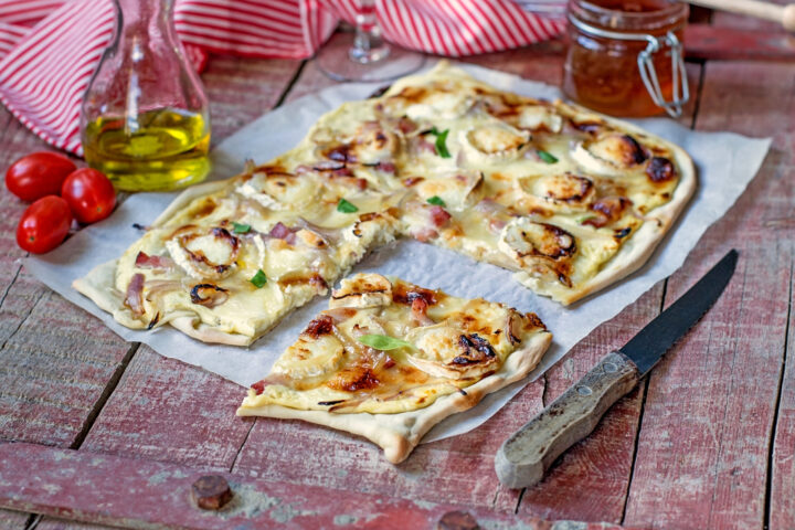 Eine rechteckige WASGAU Fladenbrotpizza mit geschmolzenem Käse, Zwiebeln, grünem Paprika und gebräuntem Belag liegt auf Pergamentpapier. Eine Scheibe ist ausgeschnitten. Auf dem rustikalen Holztisch liegen Traubentomaten, ein Glas mit Öl, Soße und ein Messer.