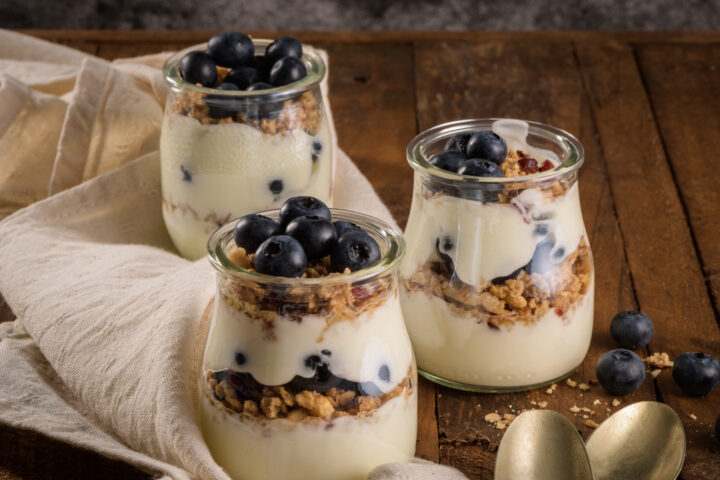 Drei mit WASGAU Joghurt, Müsli und frischen Blaubeeren gefüllte Gläser stehen auf einem rustikalen Holztisch, daneben ein beiges Tuch und zwei goldene Löffel. Einige lose Blaubeeren sind in der Nähe verstreut.