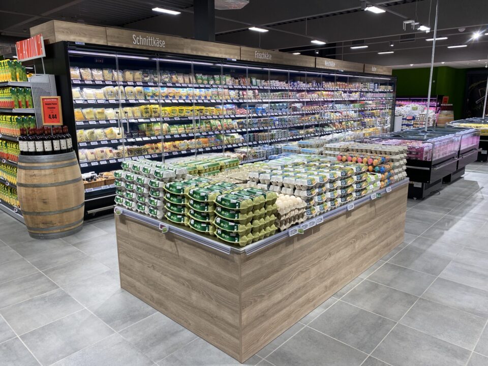 In einem WASGAU Lebensmittelgeschäft stehen Kartons mit Eiern auf einer hölzernen Insel, umgeben von Kühlregalen mit Milchprodukten, Käse und Feinkostartikeln unter heller Deckenbeleuchtung.