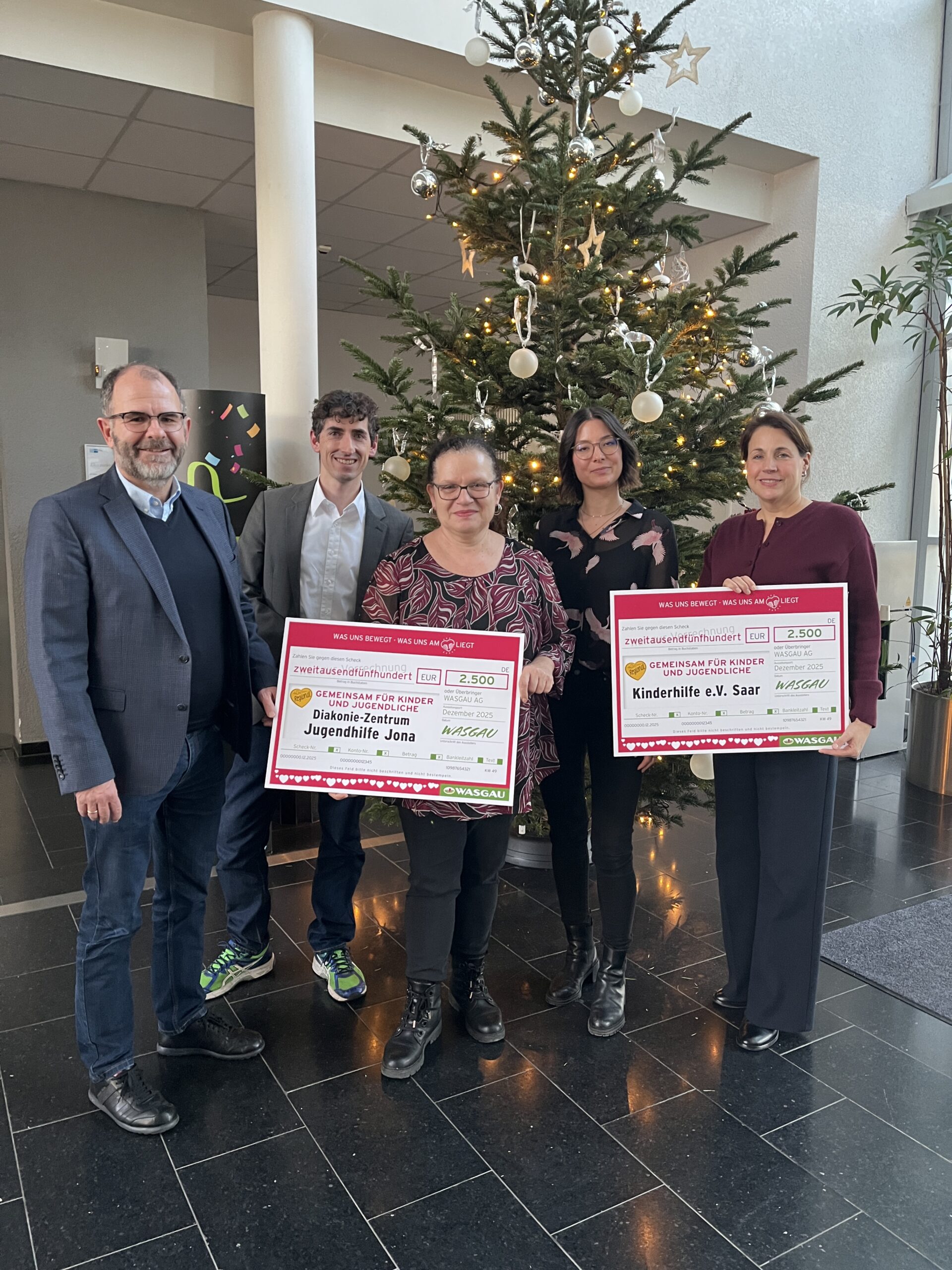 Fünf Personen stehen an einem Weihnachtsbaum, halten zwei große Spendenschecks für die KINDER Herzenswochen 2025 und lächeln in die Kamera.