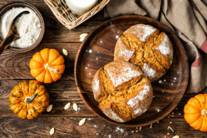 Zwei runde Kürbisbrote liegen auf einer Holzplatte, umgeben von Kürbissen, Kernen, Mehl und einem Glas Milch.