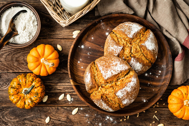 Zwei runde Kürbisbrote liegen auf einer Holzplatte, umgeben von Kürbissen, Kernen, Mehl und einem Glas Milch.