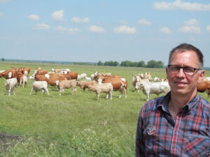 Ein Mann im karierten Hemd steht vor einer Kuhherde, die auf einer Wiese unter blauem Himmel weidet und die höchste Qualität für Premium Fleisch gewährleistet.