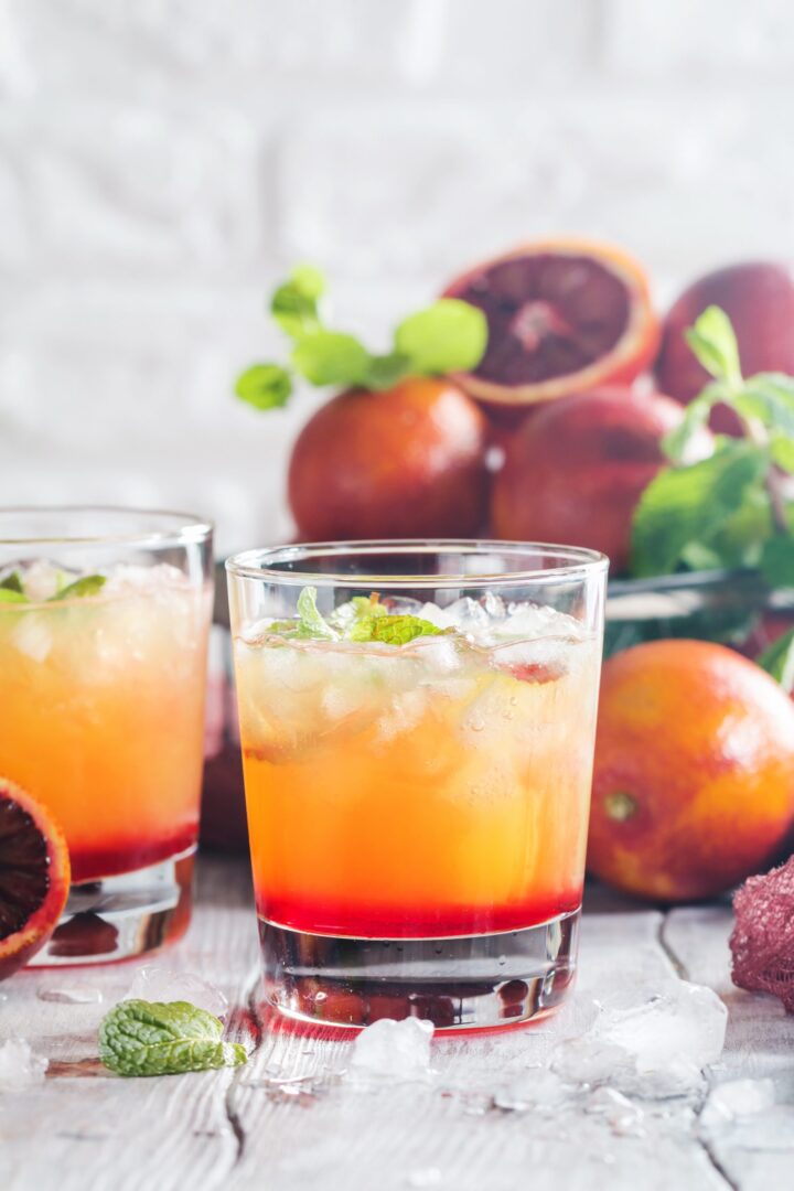 Zwei Gläser mit einem bunten Zitruscocktail mit zerstoßenem Eis, garniert mit Minze, stehen auf einer weißen Holzfläche. Frische Blutorangen und Minzblätter von WASGAU sind im Hintergrund verstreut.