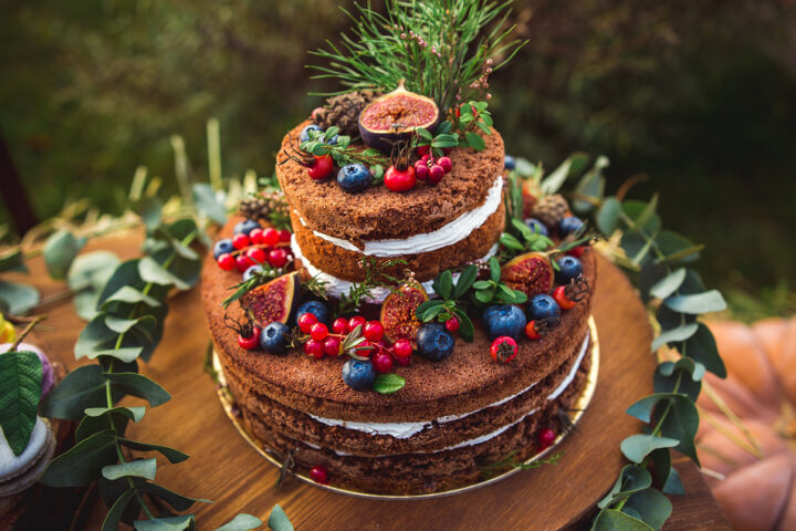 Zweistöckiger nackter Kuchen mit Beeren, Feigen, Tannenzweigen und Grünzeug auf einem Holzbrett - perfekt für eine festliche Tafel neben einer traditionellen Weihnachtspyramide.