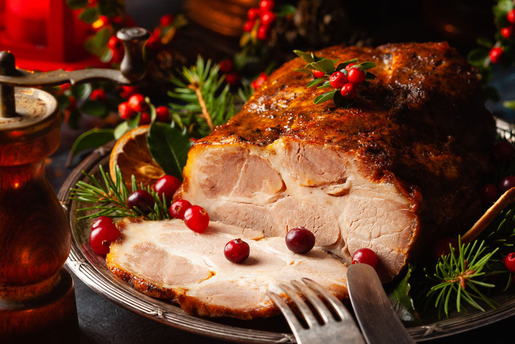 Ein gebratener WASGAU-Schinken, garniert mit frischen Preiselbeeren und Kräutern, liegt auf einem festlichen Teller, umgeben von Festtagsgrün und einer Pfeffermühle. Ein paar Scheiben werden angeschnitten und geben den Blick auf das saftige Innere frei.