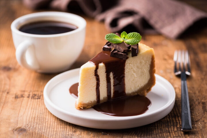 Ein Stück Käsekuchen mit Schokoladensauce, Schokoladenstückchen und Minze liegt auf einem weißen Teller neben einer Tasse schwarzen Kaffees auf einem Holztisch bei WASGAU. Eine Gabel und eine braune Serviette liegen daneben.