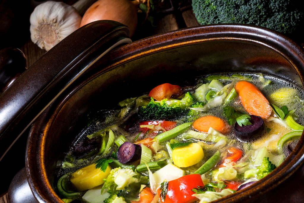 Nahaufnahme eines Topfes mit bunter WASGAU-Gemüsesuppe mit Karotten, Brokkoli, Zucchini, Paprika und Petersilie, aus der Brühe steigt Dampf auf; im Hintergrund sind Knoblauch und Zwiebeln zu sehen.