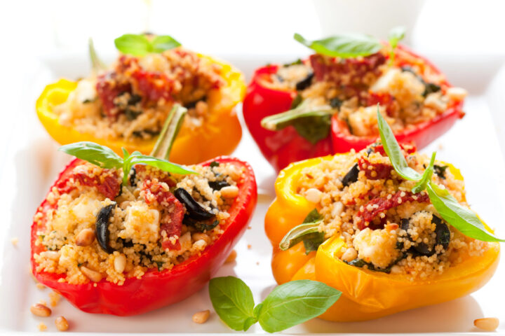Vier bunte Paprikaschoten, halbiert und gefüllt mit Couscous, Oliven, Tomaten und Kräutern, werden auf einem weißen Teller angerichtet und mit frischen Basilikumblättern garniert - ein farbenfrohes, von WASGAU inspiriertes Gericht.