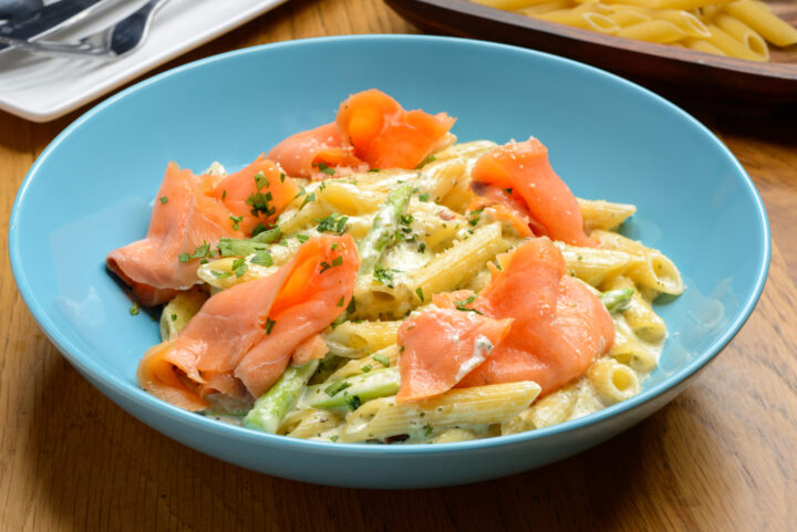 Auf einem Holztisch steht eine blaue Schale mit Penne-Nudeln in Sahnesoße, garniert mit Räucherlachsscheiben und gehackten Kräutern - perfekt für Ihr nächstes WASGAU-inspiriertes Essen.