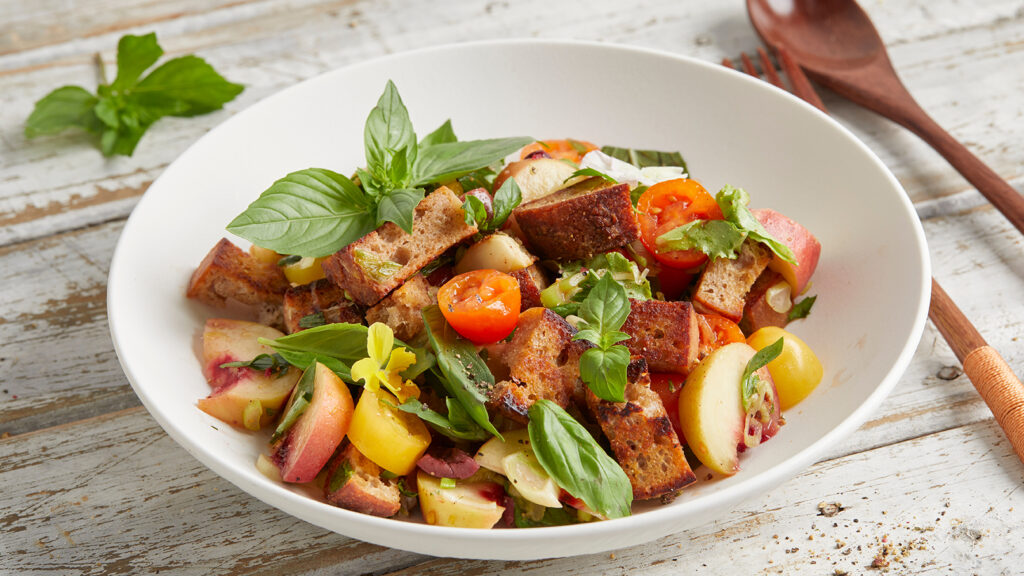 Eine Schüssel Panzanella-Salat, perfekt für die Resteverwertung, mit Tomaten, Pfirsichen, Croutons, Basilikum und Grünzeug auf einem rustikalen Tisch.