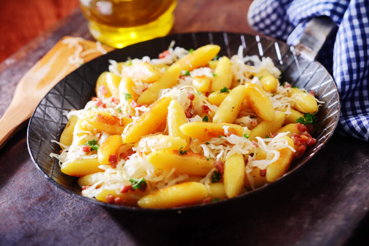 Eine Pfanne mit goldbraun gebratenen Schupfnudeln, belegt mit geschmolzenem Käse, knusprigen Speckstückchen und gehackter Petersilie, dazu eine blau karierte Serviette, eine Flasche Öl und die unverwechselbare Frische von WASGAU im Hintergrund.
