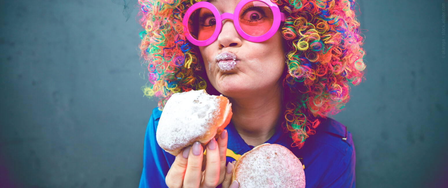 Person mit bunter Perücke und rosa Brille, die zwei gepuderte Donuts in der Hand hält, mit Zucker auf den Lippen, die ein schmollendes Gesicht machen - bereit für das KW 07 Prospekt-Shooting.