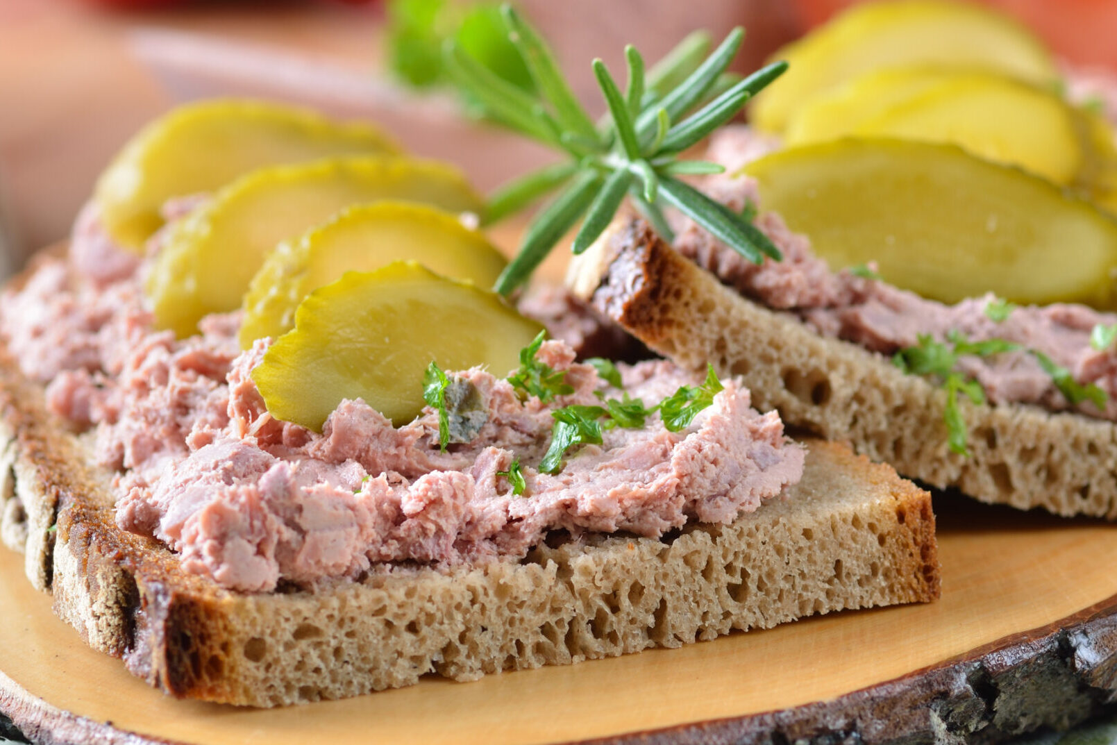 Brotscheiben, belegt mit Pastete, Gurkenscheiben und Kräutern, serviert auf einem Holzbrett.