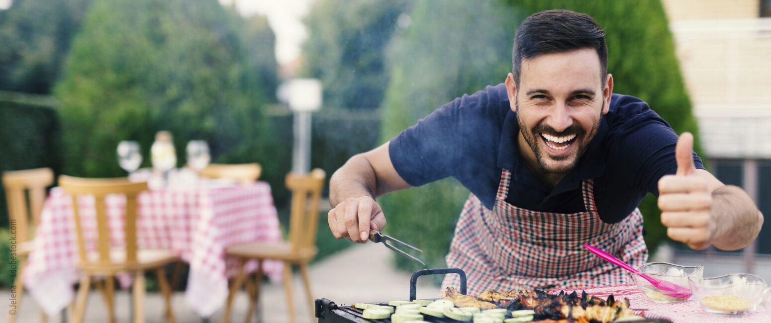 Mann mit dunklen Haaren, dunklem Bart und Schürze steht in einem Garten, beugt sich über einen Grill mit einer Grillgabel in der einen Hand, mit der anderen Hand zeigt er ein Daumen nach oben und lächelt frontal in die Kamera. Auf dem Grill liegt verschiedenes Grillgut wie Spieße, Gemüse und Fisch.