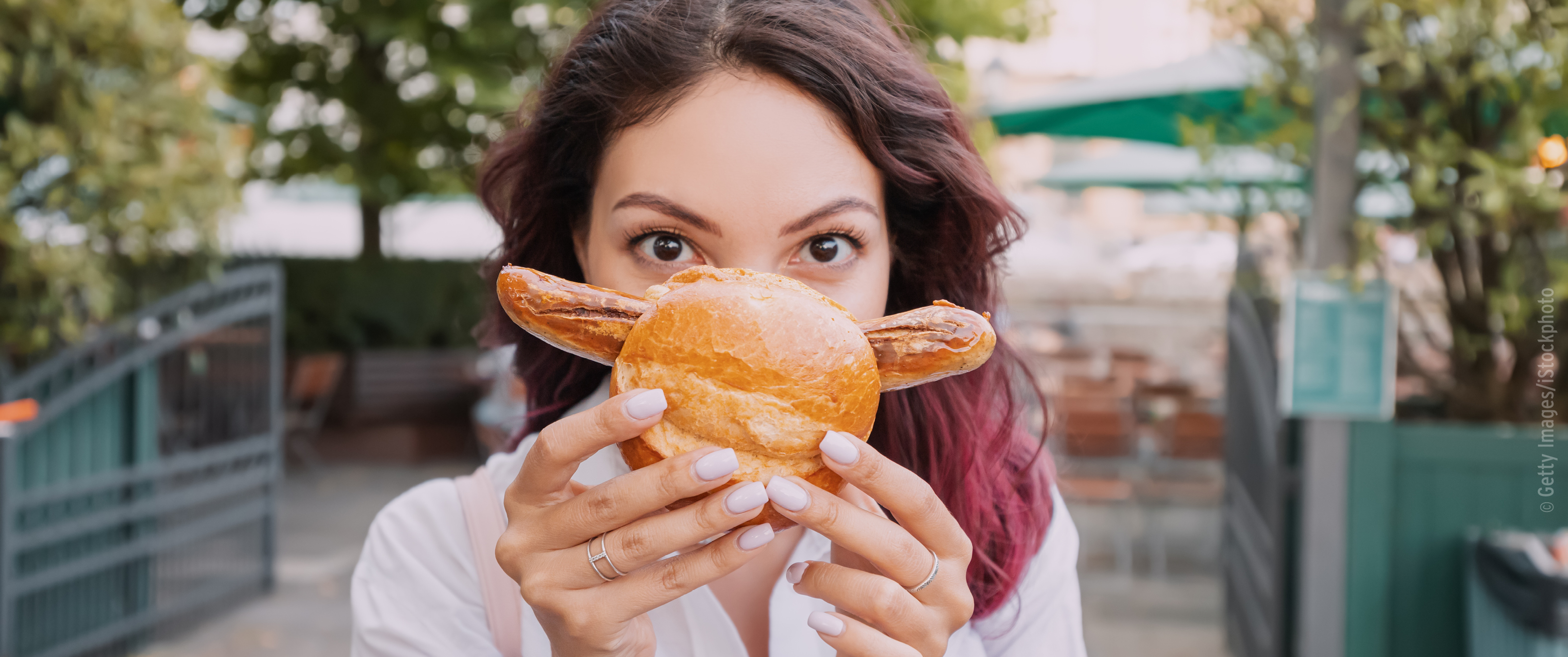 Eine Frau in einem Café oder Park hält spielerisch ein Brezelbrötchen in Form eines Gesichts vor sich und fängt damit einen lustigen Prospekt-Moment im Freien ein. KW 32.