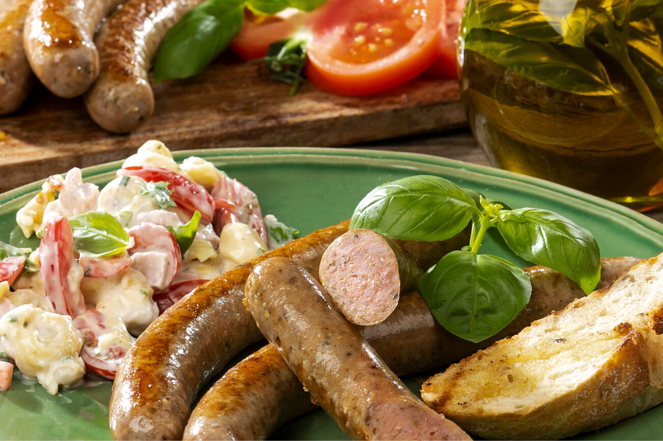 Gegrillte Würstchen mit einer Scheibe Brot, Salat und Basilikum auf einem grünen Teller, mit Tomaten im Hintergrund.