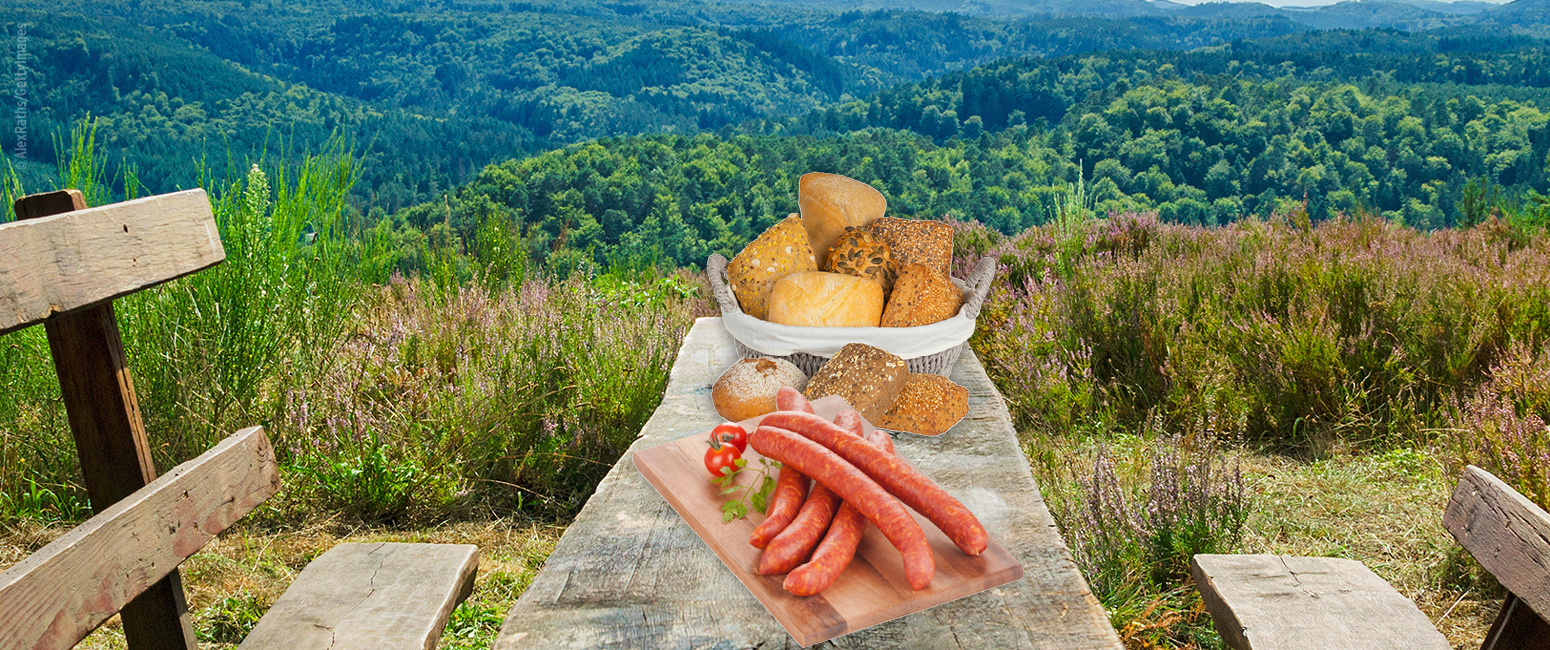 Ein rustikaler Tisch im Freien mit Würstchen, Brot und Brötchen aus der KW 36 Prospekt, mit Blick auf eine grüne, bewaldete Berglandschaft.