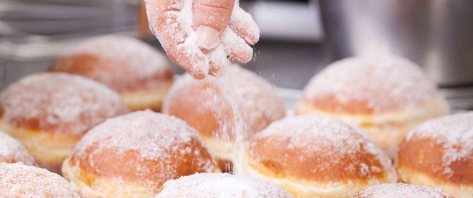 Eine Hand streut Zucker auf frisch gebackene, runde Donuts, die auf einem Tablett angeordnet sind - perfekt für Ihren KW 39 Prospekt.