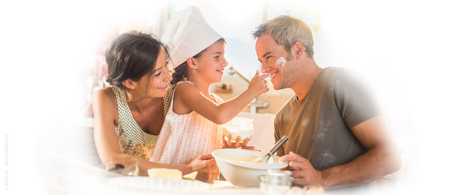 Ein lächelndes Mädchen mit Kochmütze, abgebildet in KW 45 Prospekt, streut spielerisch Mehl auf die Nase ihres Vaters, während ihre Mutter in der Küche zusieht.