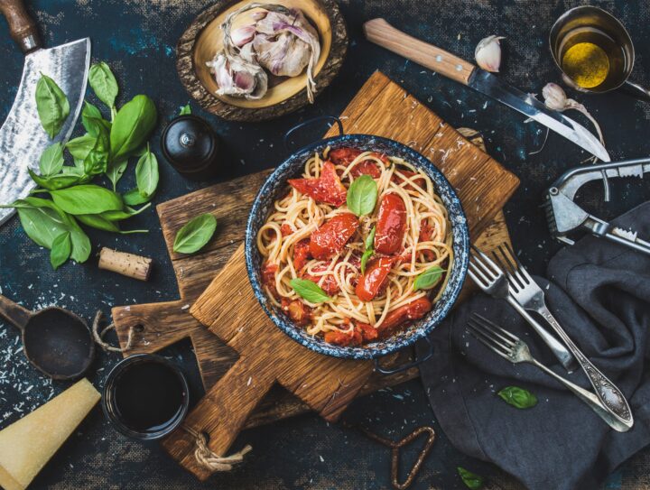 Eine Schüssel Spaghetti mit Tomatensauce und Basilikum steht auf einem Holzbrett, umgeben von Gabeln, Knoblauch, Olivenöl, einem Weinglas, geriebenem Käse und frischen WASGAU Basilikumblättern auf einem rustikalen, dunklen Hintergrund.