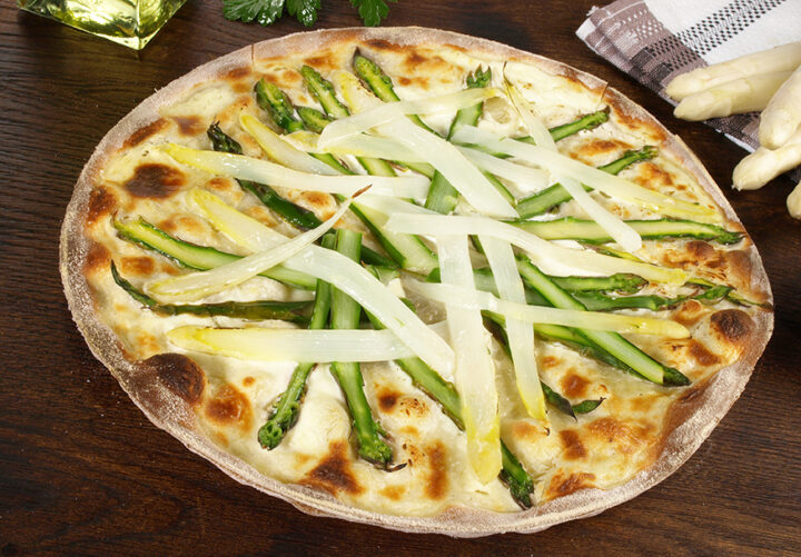 Von Flammkuchen inspirierte Pizza, belegt mit weißem und grünem Spargel, auf einem Holztisch neben einem Handtuch und frischem rohen Spargel.