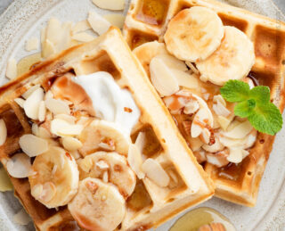 Zwei Waffeln mit Bananenscheiben, Mandelblättchen und einem Spritzer Honig auf einem Teller. Neben den Waffeln befinden sich ein Honiglöffel und eine Minzgarnitur. Der Text lautet: 
