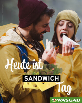 Ein glückliches Paar in gelben Regenmänteln genießt ein leckeres Sandwich im Freien, wobei der deutsche Text und das Wasgau-Logo sichtbar sind.