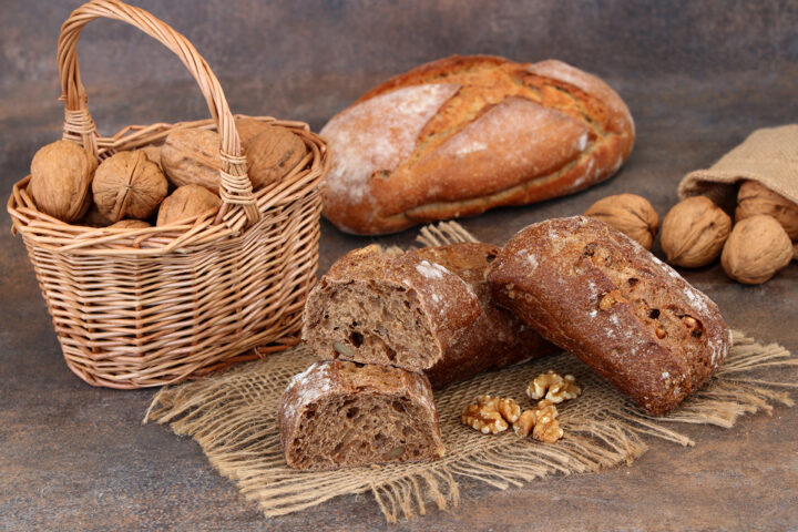Ein Korb mit Walnüssen und geschnittenem Vollkornbrot auf Sackleinen, mit Walnussbrötchen, zusätzlichen Walnüssen und einem Laib im Hintergrund.