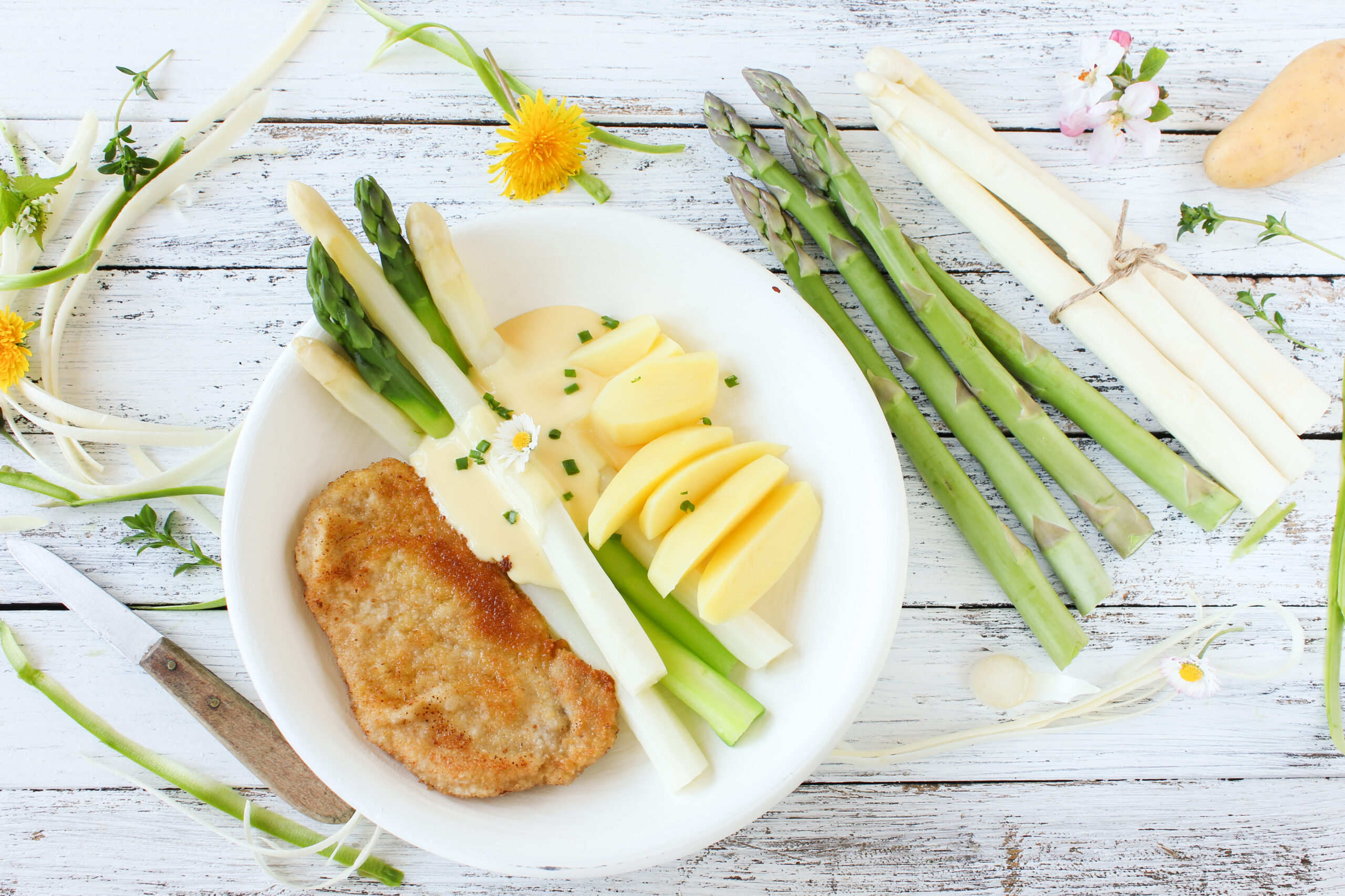 Teller mit Schnitzel, weißem und grünem Spargel, Kartoffeln und Soße, auf einem rustikalen weißen Holztisch.