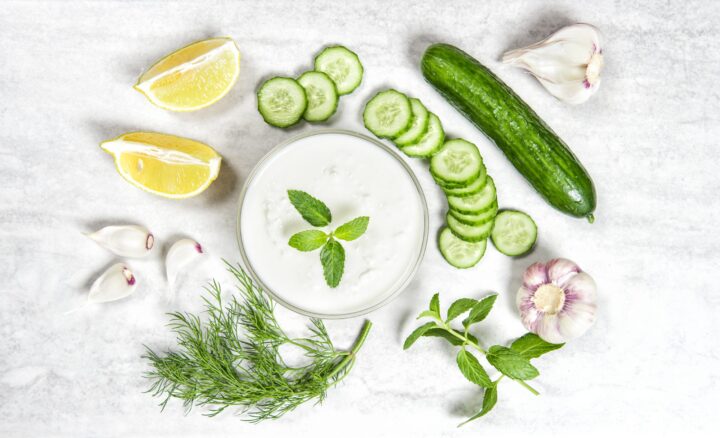 Eine Schale mit WASGAU-Joghurtdip, garniert mit Minzblättern, umgeben von Gurkenscheiben, Zitronenspalten, ganzen Knoblauchzehen, Dill und frischer Minze auf einer weißen Marmorplatte.