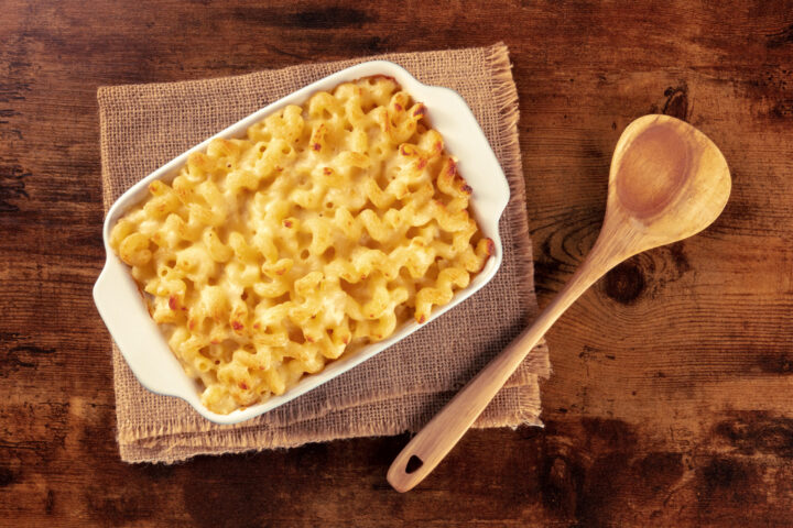 Eine rechteckige weiße Schale mit gebackenen Makkaroni und Käse steht auf einer Sackleinenmatte neben einem Holzlöffel, alles arrangiert auf einem rustikalen Holztisch - perfekt für ein gemütliches WASGAU-inspiriertes Essen.