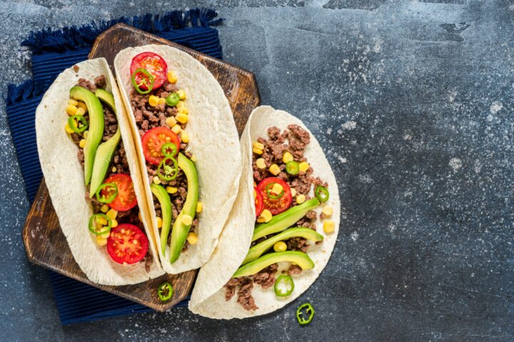 Drei weiche Taco-Schalen, gefüllt mit Rinderhackfleisch, geschnittener Avocado, halbierten Kirschtomaten, Mais und Jalapeño-Scheiben, werden auf einem Holzbrett und blauem Tuch präsentiert - klassische Aromen, die mit frischen WASGAU Zutaten vor einem dunkel strukturierten Hintergrund hervorgehoben werden.