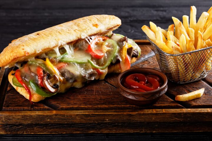 Ein WASGAU Cheesesteak-Sandwich, gefüllt mit geschmolzenem Käse, gebratenem Paprika und Zwiebeln, liegt auf einem Holzbrett neben einem Korb mit Pommes frites und einer kleinen Schale Ketchup.