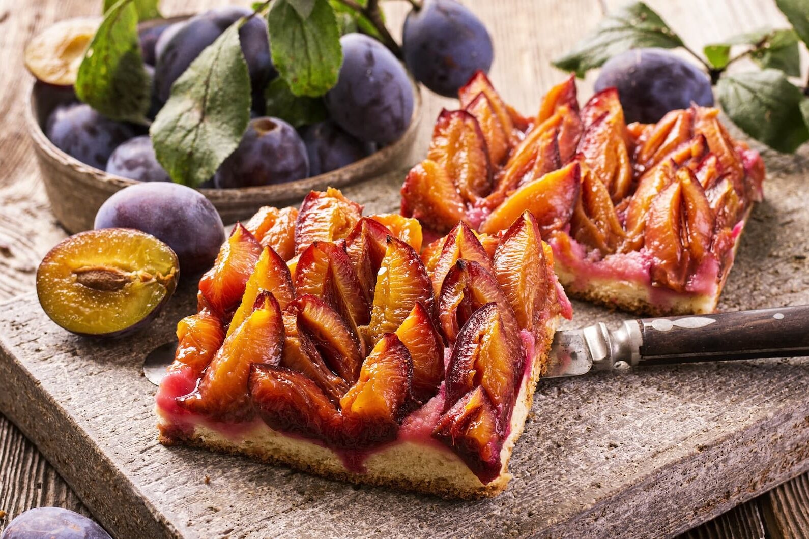 Zwei Scheiben Pflaumenkuchen auf einer Steinplatte mit frischen Pflaumen und Blättern im Hintergrund.